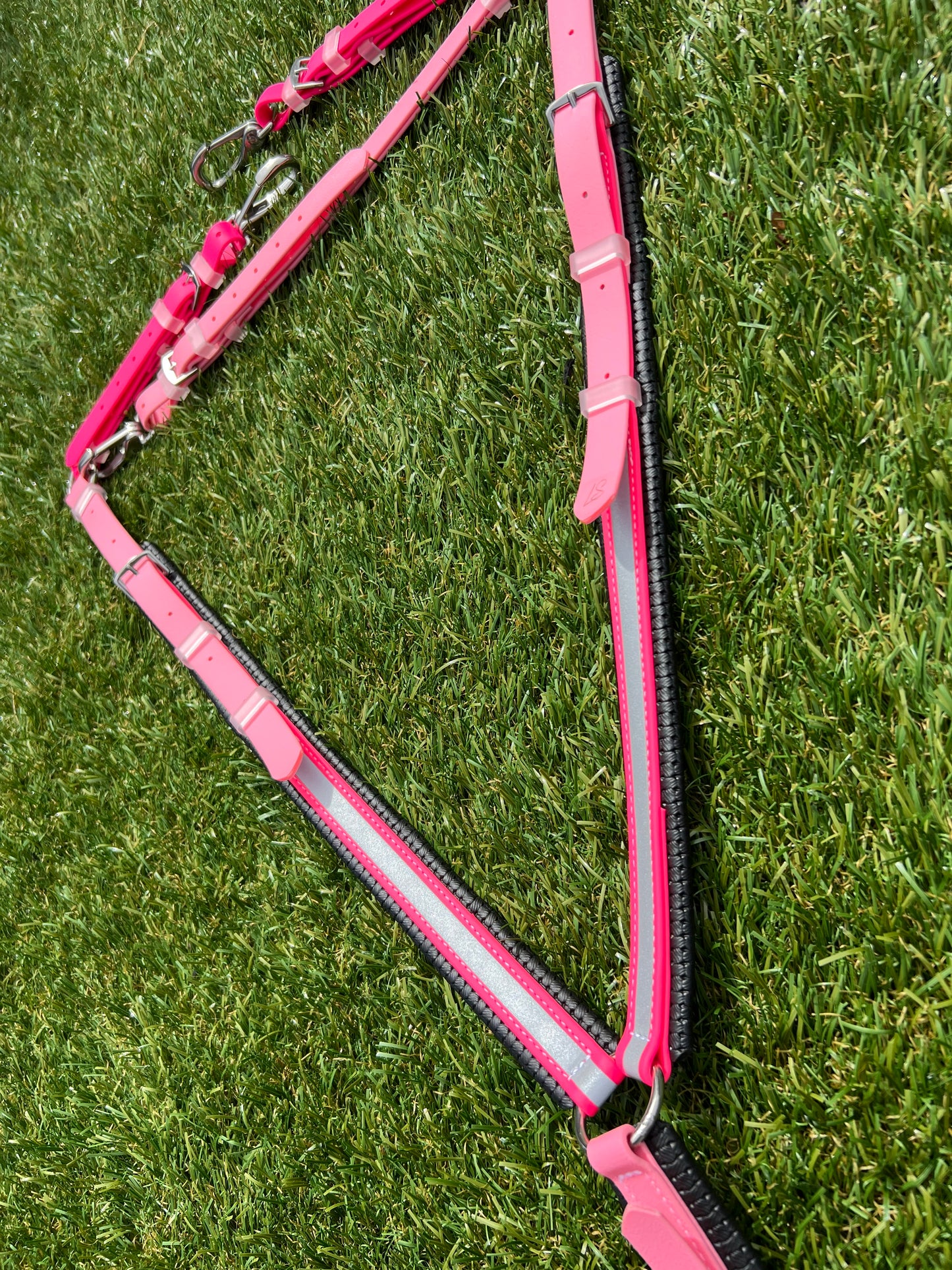 A pink LS Equestrian Customisable Reflective BioThane® Breastplate with adjustable buckles and reflective stripes rests on a grassy surface. Durable, safe, and high-quality horse tack.