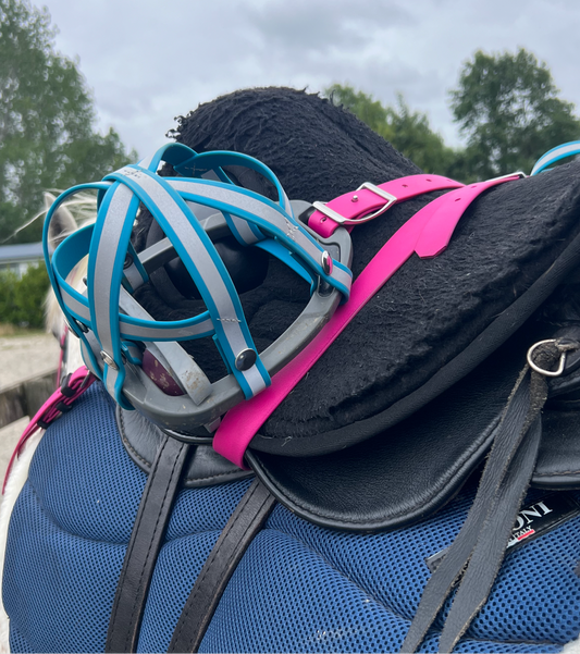 A close-up showcases a blue bag adorned with a black and pink shoulder strap, topped by a black sweater or jacket. Atop the bag lies the LS Equestrian "Design Your Own - Reflective BioThane®️ Snap Smart Stirrup Cages," featuring a blue and gray design reminiscent of sports or safety equipment. In the background, trees stand beneath a cloudy sky.