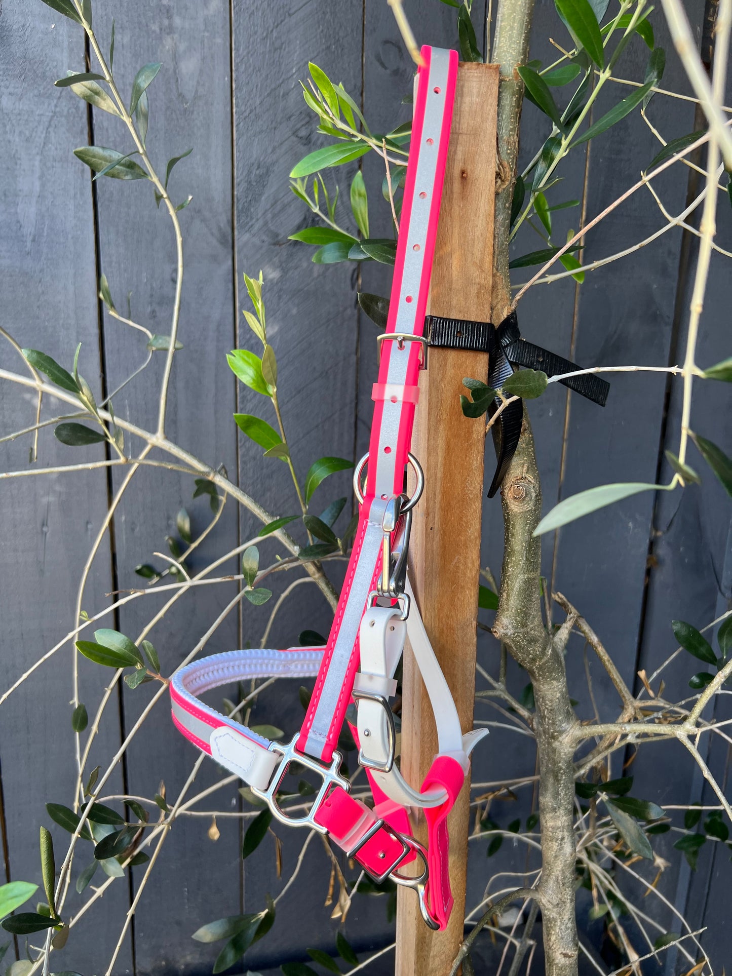 A Reflective BioThane®️ Halter - Padded design your own, from LS Equestrian, in neon pink and light gray hangs on a wooden post next to a leafy plant. Its reflective material contrasts with the dark wooden fence in the background, ensuring both visibility and style for night riders.