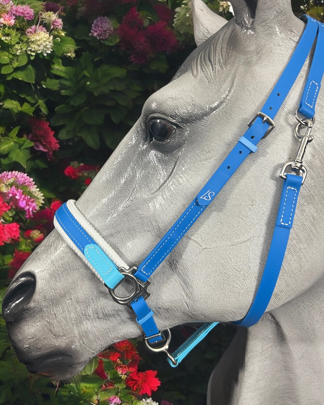 A model horse head displays an eye-catching blue halter made with BETA® BioThane® from LS Equestrian's Side Pull Bitless Bridle Option 1 - Design Your Own, beautifully framed by vivid pink and purple flowers and rich green leaves.