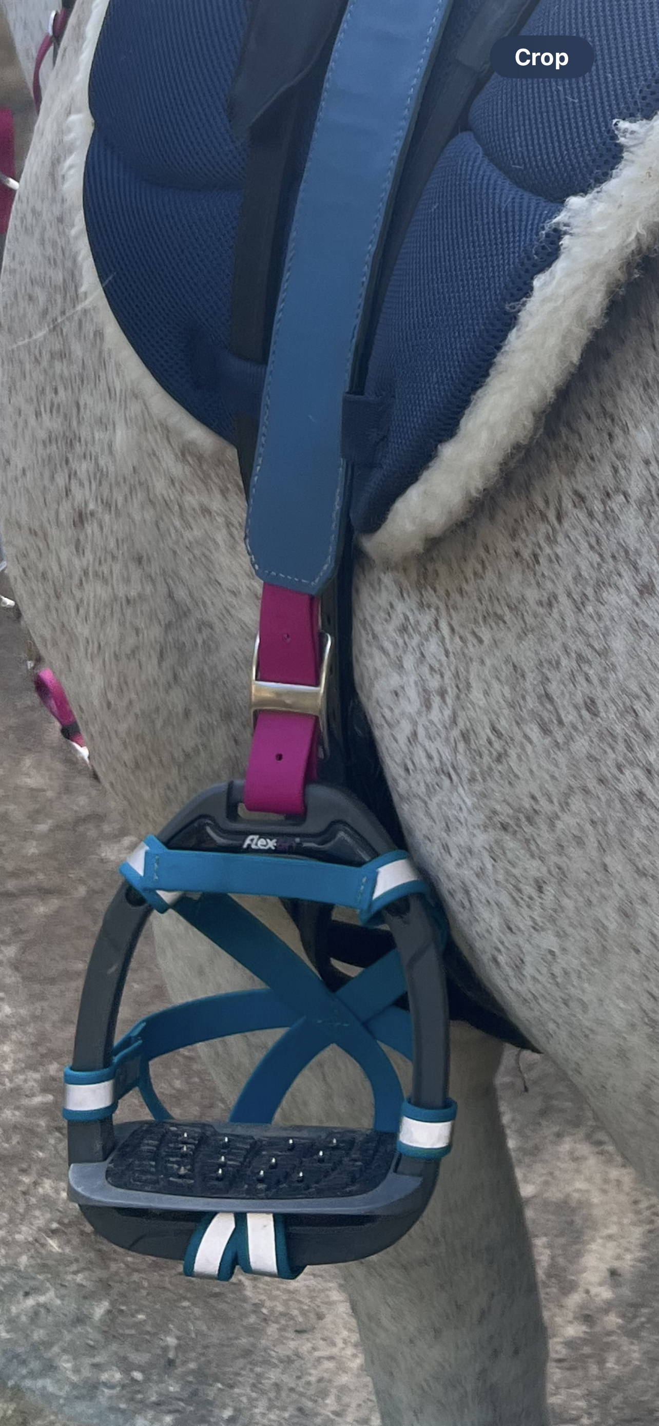 A detailed view of a horse saddle showcases LS Equestrian's Design Your Own - Reflective BioThane®️ Snap Smart Stirrup Cages, highlighted by their blue and black straps. The saddle boasts a striking black and blue pattern crafted from Beta BioThane Colours, complemented by a sheepskin lining underneath. The horse's light gray speckled coat is partially visible in the background.
