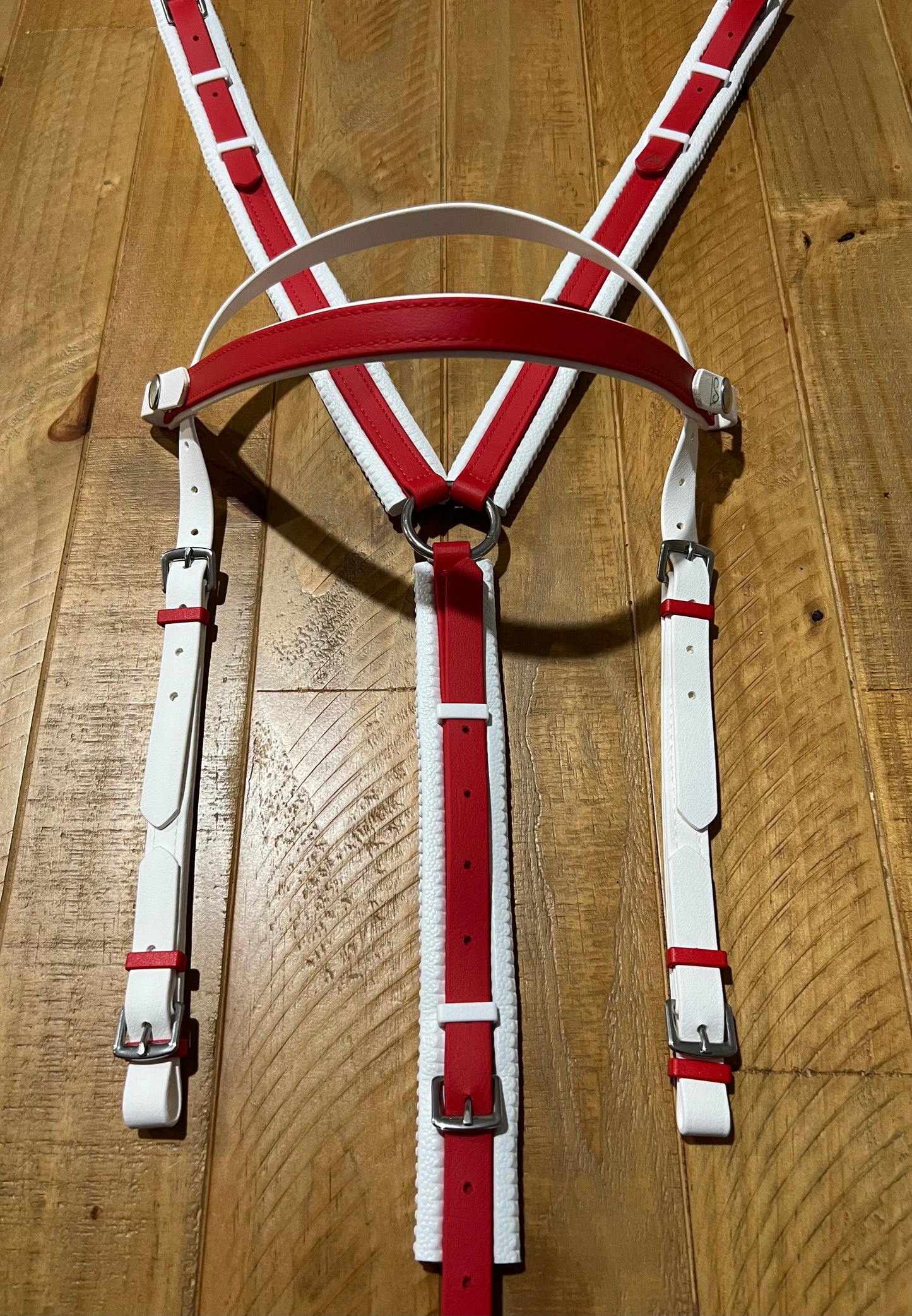 An LS Deux Endurance Headstall by LS Equestrian, featuring a red and white leather harness with adjustable straps and metal buckles, reminiscent of endurance riders' gear, is displayed flat on a wooden surface.