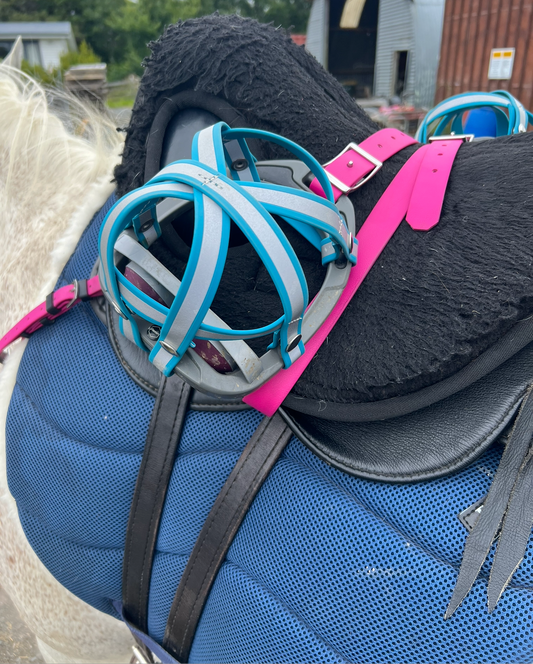 A blue and black horse saddle featuring a black cushion is complemented by LS Equestrian's pink BioThane®️ Stirrup Cages from their "Design Your Own - Replacement Snap Smart" collection. Resting atop the saddle is a set of blue spherical safety stirrups. The rustic charm of the scene is enhanced by a background that showcases a rusted barn and lush greenery.