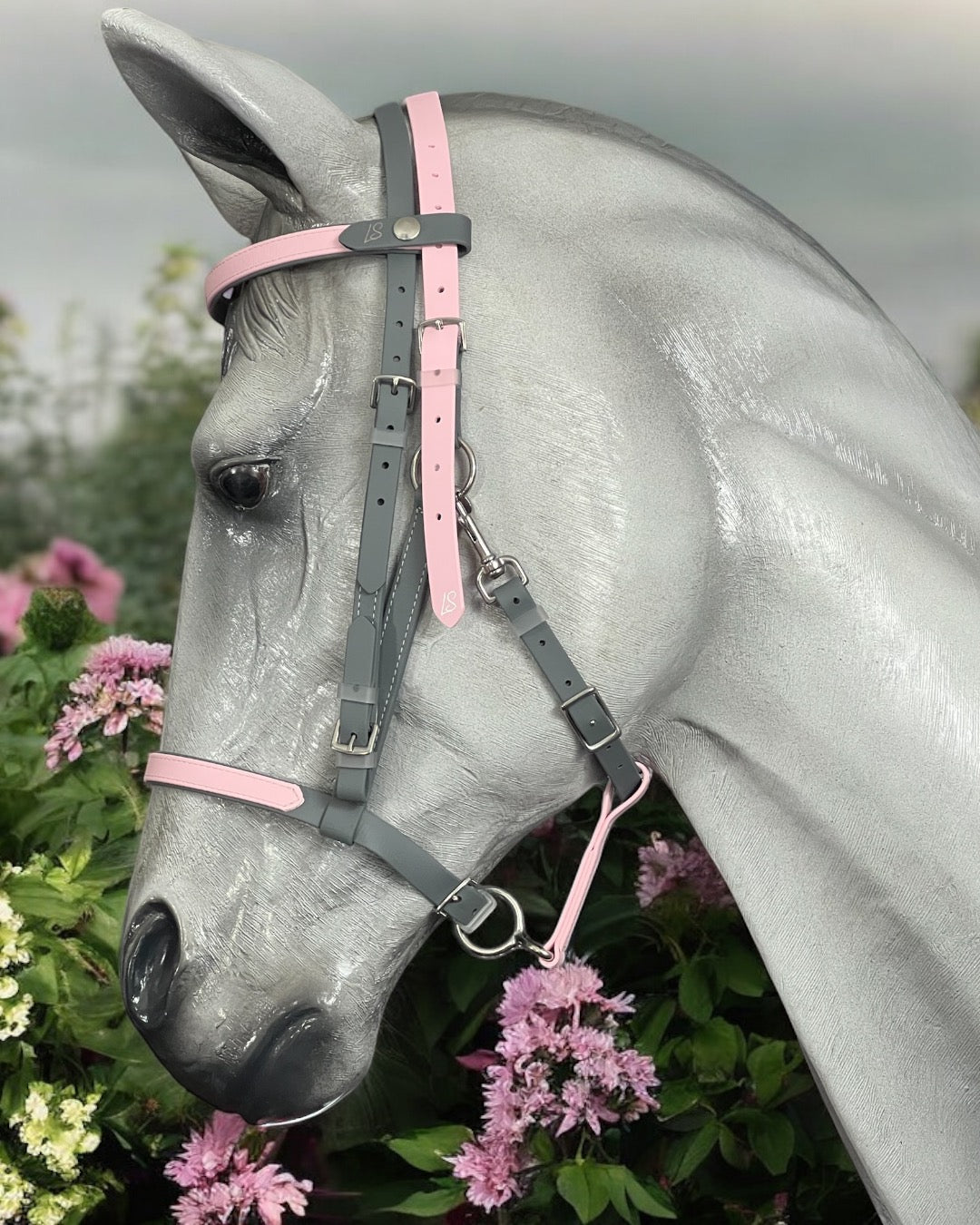 A gray horse is wearing an LS Equestrian LS Deux Lightweight 2-Part Endurance Bridle with pink accents, standing near blossoming pink flowers against a soft, overcast sky.