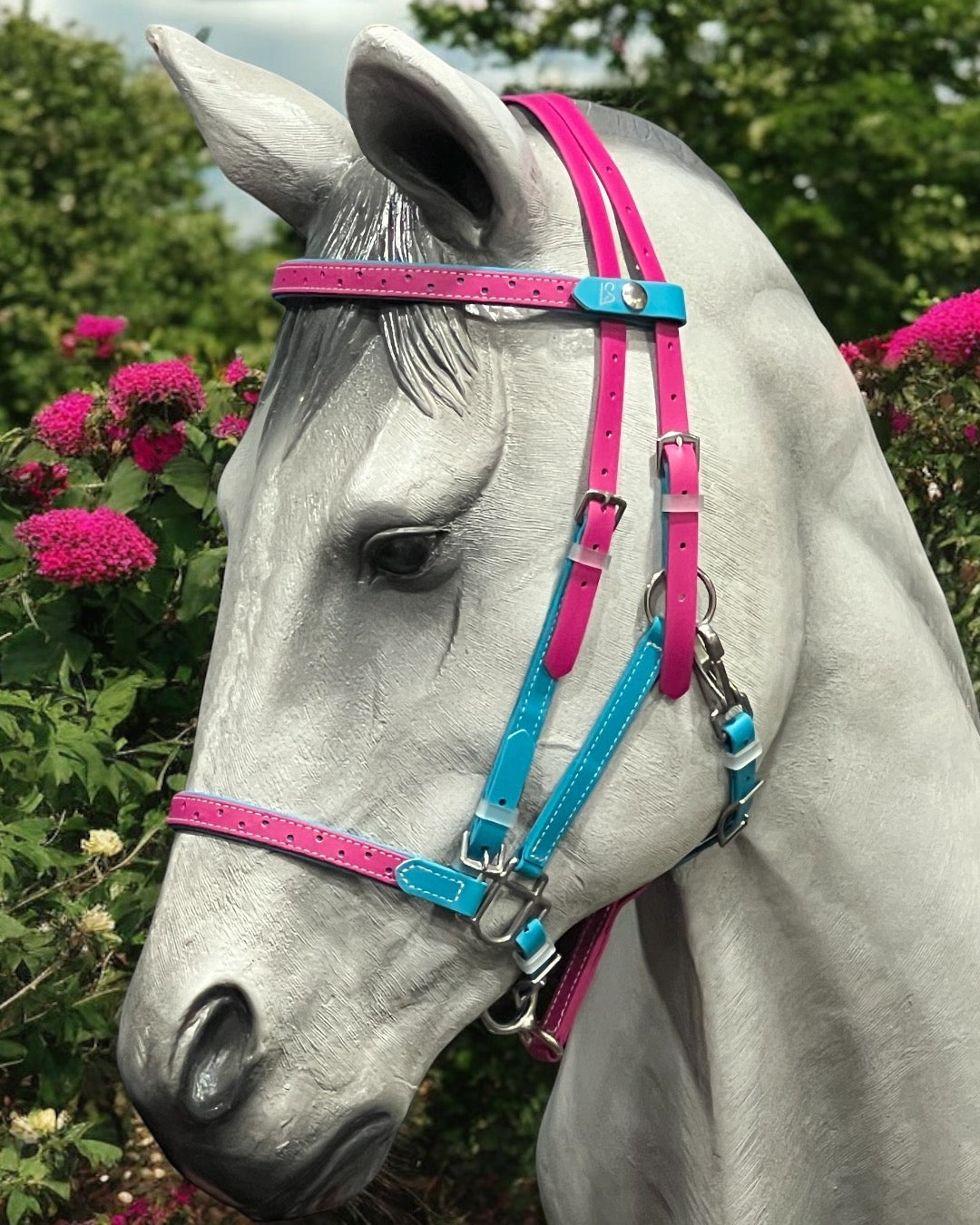 A white horse wears the LS Equestrian LS Deux Endurance 2-Part Detailed Bridle, featuring vibrant pink and blue Beta BioThane straps. Bright pink flowers blend with lush greenery in the background, highlighting handcrafted excellence.