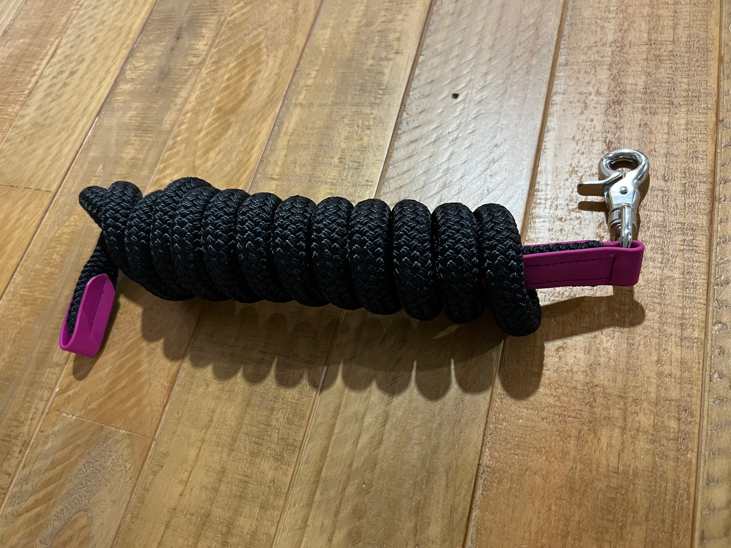 An LS Equestrian "LS Lead Rope - Design your Own" featuring a coiled black rope, stainless steel clip, and pink handle is resting on a wooden floor.