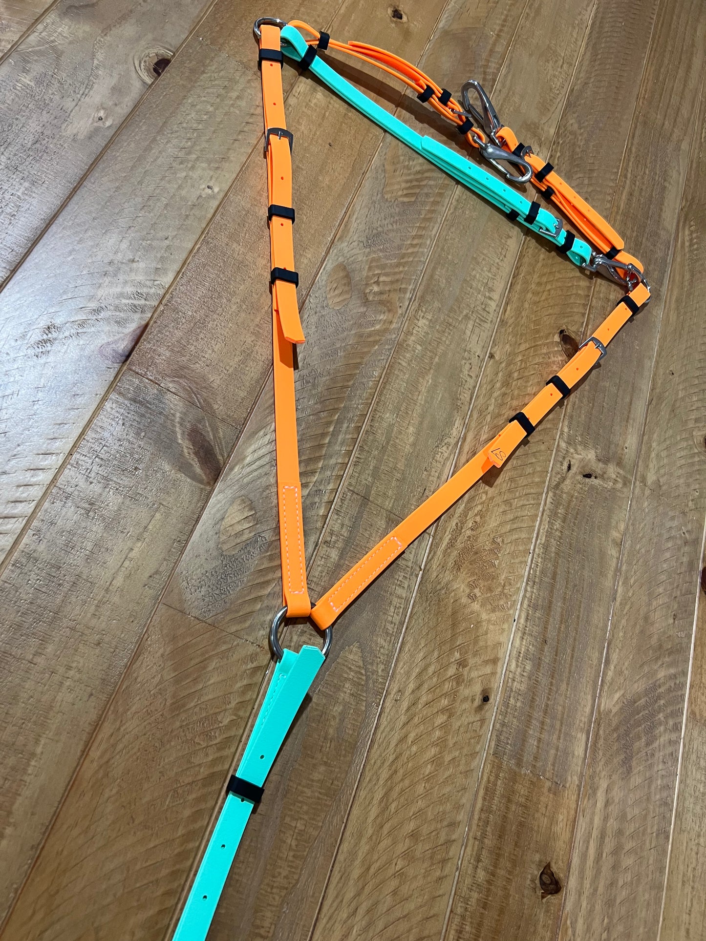 A Breastplate Zero Padding - Design your Own from LS Equestrian, crafted with custom-made orange and turquoise straps, is laid out on a wooden floor. The breastplate includes stainless steel fittings such as metal rings and buckles for adjustments.