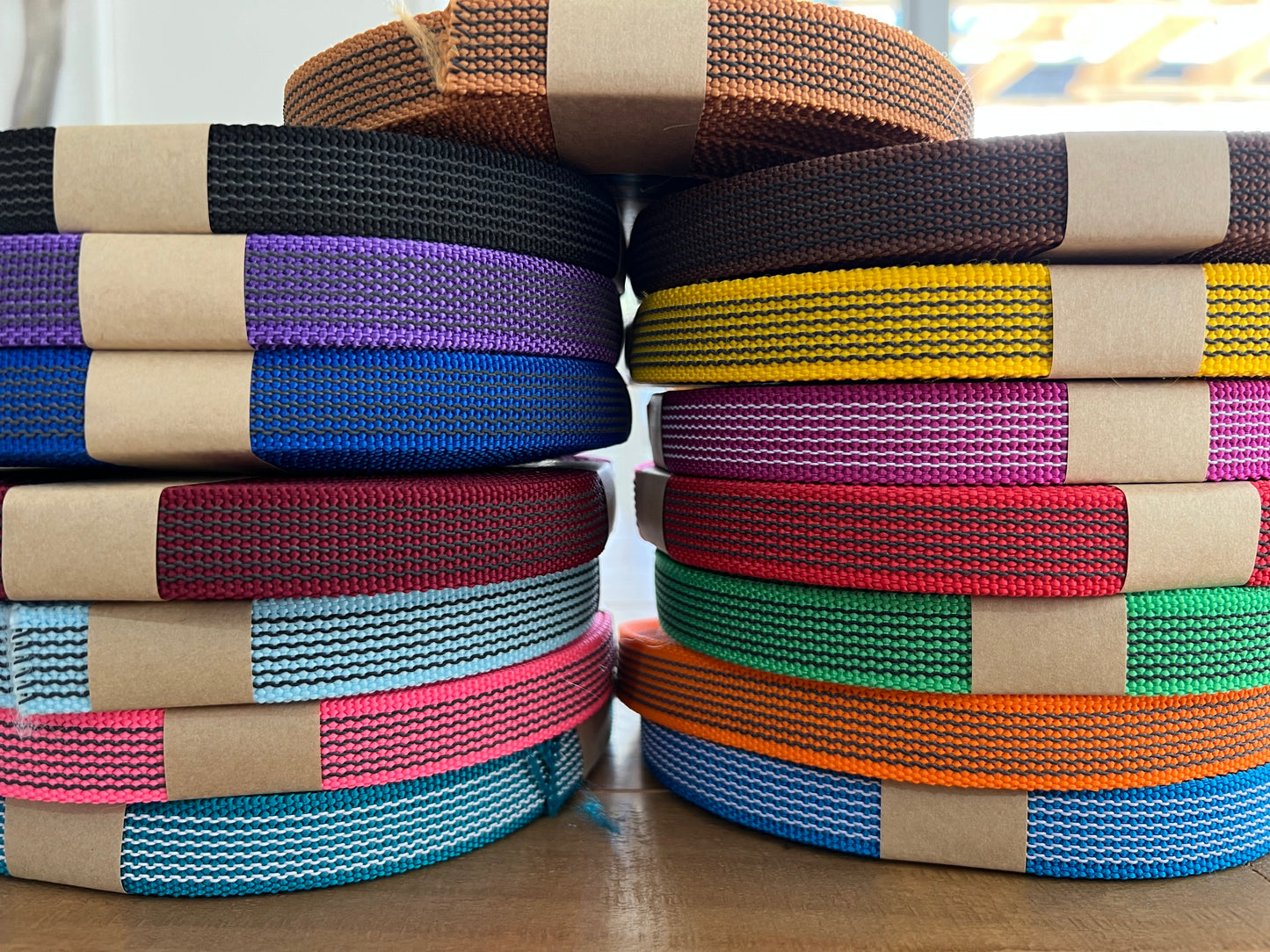 Two stacks of LS Equestrian's "Webbed Reins - Design Your Own" sit side by side on a table, showcasing an array of vibrant colors such as purple, yellow, pink, and blue. Each roll is tightly secured with brown paper bands, providing a firm hold on the vivid assortment.