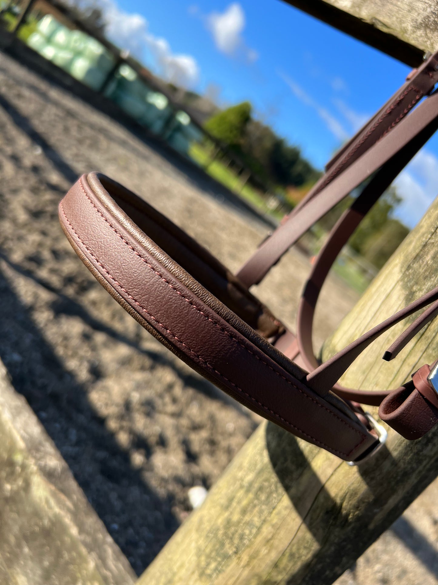 Close-up of an LS Equestrian Cavesson Bridle in brown, crafted from durable Beta BioThane, hanging on a wooden post with a sandy riding arena and blue sky creating a serene outdoor backdrop.