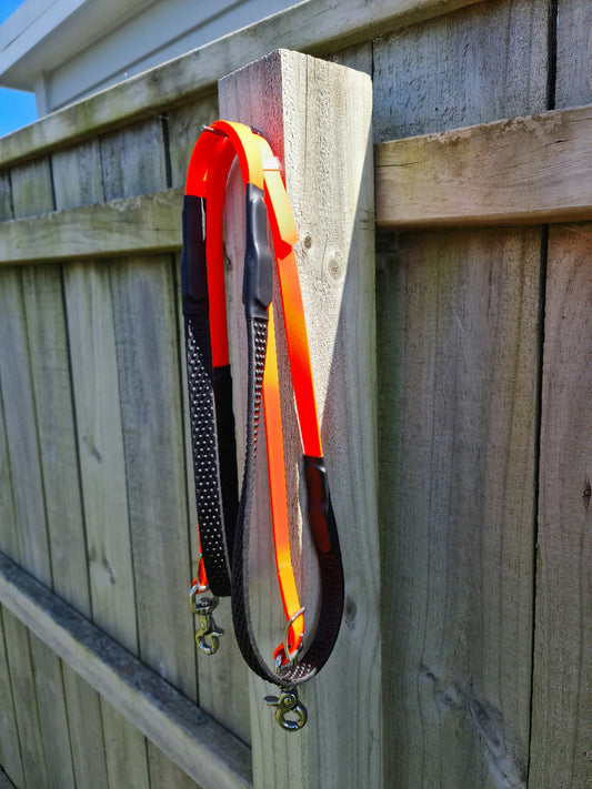 A vibrant fluro orange and black soft grip rein from the LS Equestrian collection hangs on a wooden fence under a clear blue sky.