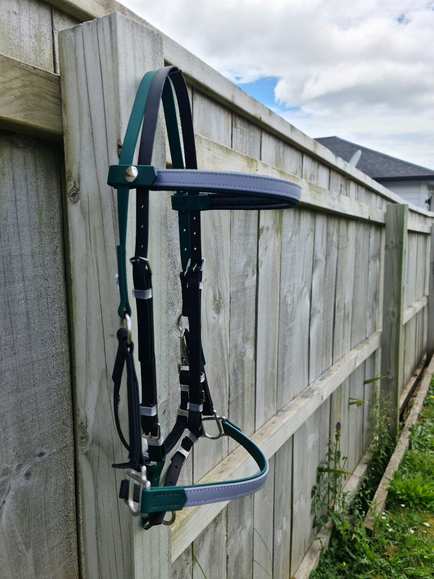 An LS Equestrian LS Deux Bridle in Hunter Green, Black, and Grey hangs on a wooden fence. The scene is outdoors, with a cloudy sky overhead and a grassy area beneath—an ideal setting for those passionate about endurance riding.
