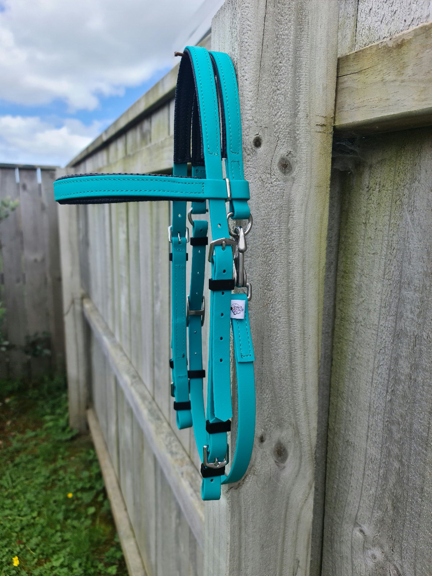A teal Padded Race Bridle from LS Equestrian hangs gracefully over a wooden fence on a cloudy day, surrounded by lush grass and foliage.