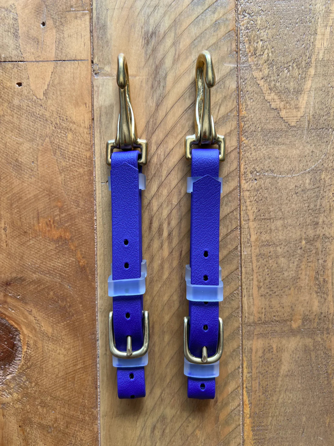 On a wooden surface, two identical LS Bit Hangers from LS Equestrian in purple leather, featuring brass hooks and buckles, are placed parallel. These straps offer adjustable sizing with multiple holes, ideal for those looking for versatile equipment such as multi bridles.