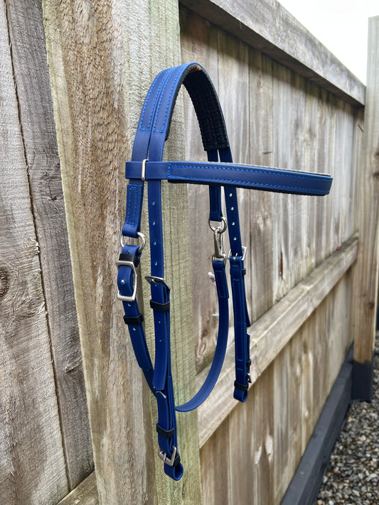 A navy Padded Race Bridle from the LS Equestrian collection hangs on a weathered wooden fence. Designed for endurance with multiple straps and buckles, the bridle features a padded section for added comfort. Gravel is scattered on the ground below.