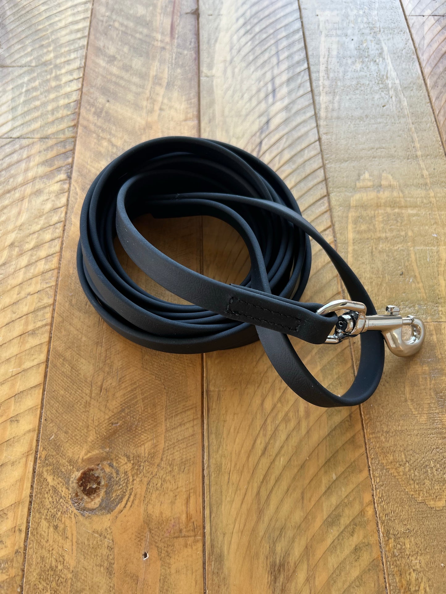 A coiled black "Training Lead - 3 metres - Design your Own" by LS Equestrian with a silver clasp lies on a textured wooden surface.