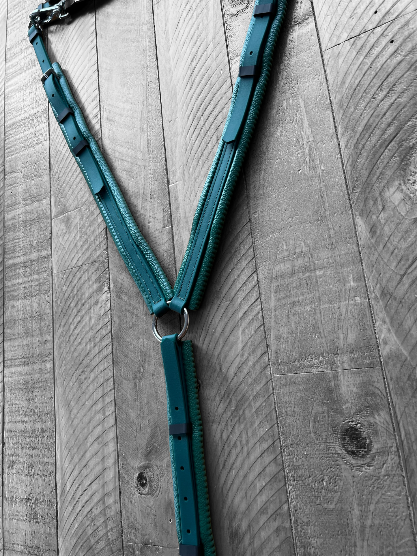 A custom-designed Padded Breastplate - Green on Green from LS Equestrian, crafted with metal rings and teal leather, is displayed on a wooden plank wall. The aged wood, featuring visible knots and grain patterns, serves as a rustic backdrop for this BioThane piece that forms a Y-shape against the textured surface.