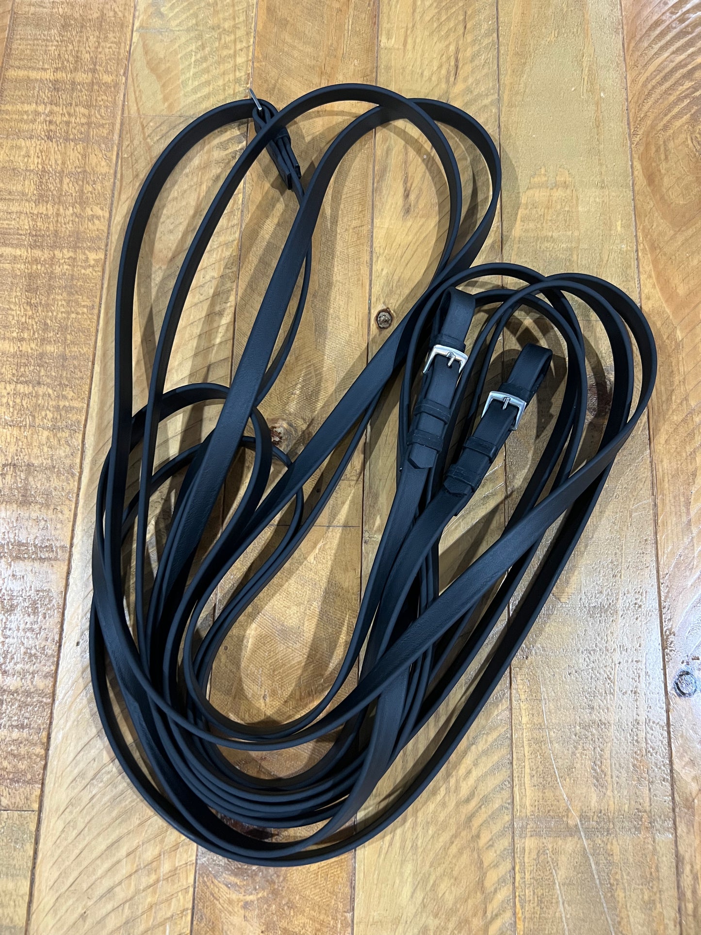 Several coiled Driving Reins - Super Heavy BioThane - Black from Ls Equestrian, featuring long leather straps with stainless steel buckles, are neatly arranged on a wooden floor that has a natural, light wood grain texture.