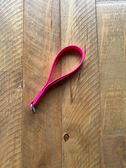 The LS Equestrian Girth Loop - Design your Own, featuring a pink rubber wristband with a metal ring, rests on a wooden surface with a herringbone pattern. The wood's natural finish highlights its grain and texture.