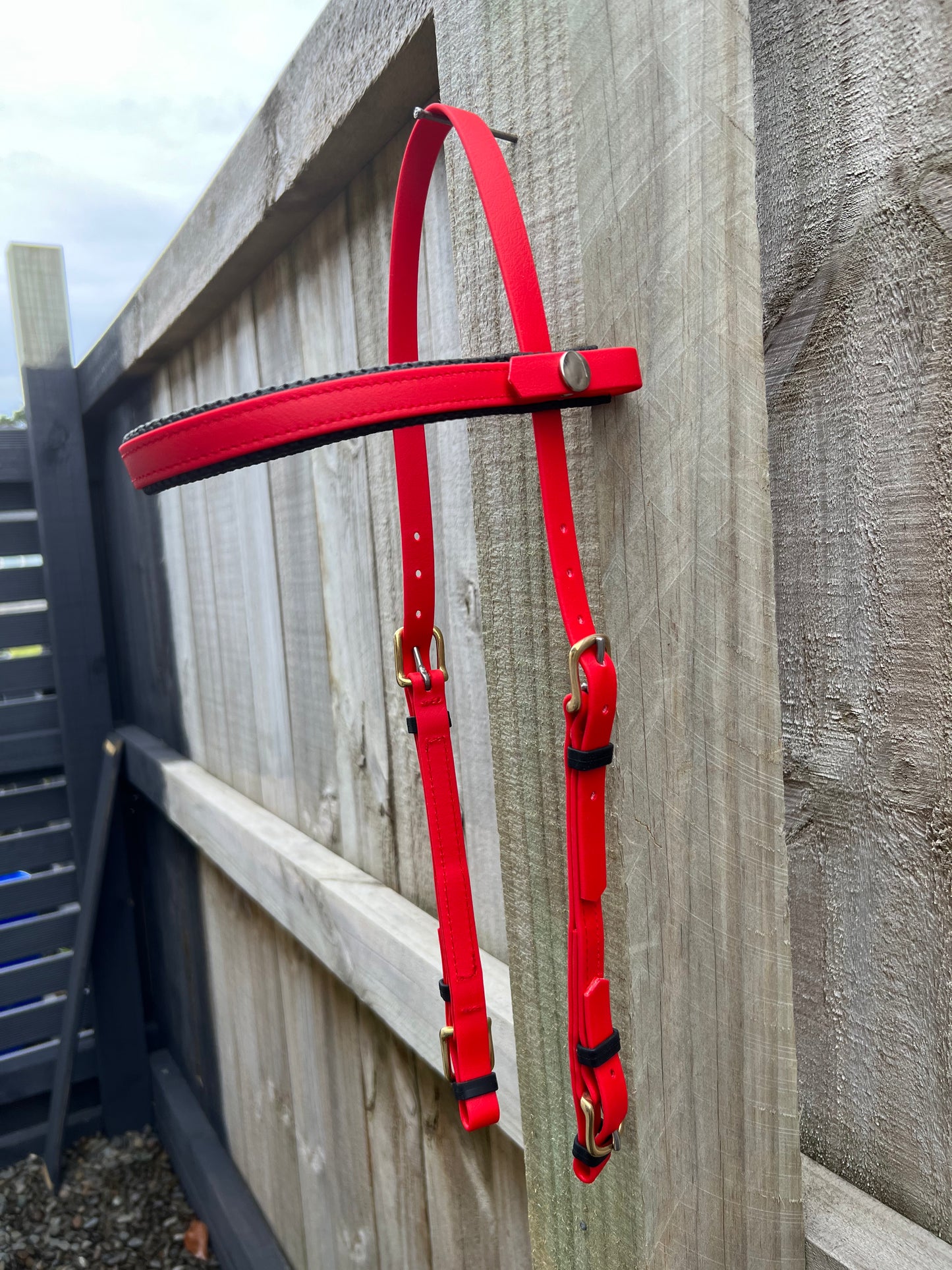 The LS Equestrian 2 Part Bridle in bright red, ideal for endurance riders, hangs on a wooden fence. It has metal buckles and a brass snap. The fence, made of horizontal wooden planks with a light natural finish, stands against a cloudy sky backdrop.