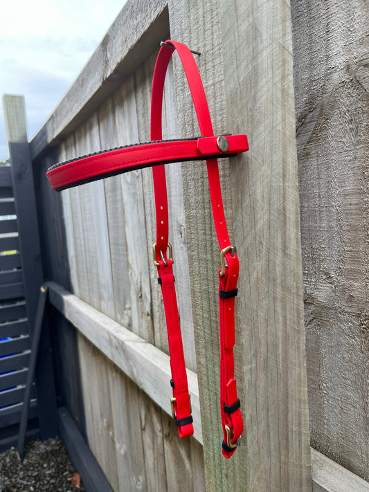 The LS Equestrian 2 Part Bridle in bright red, ideal for endurance riders, hangs on a wooden fence. It has metal buckles and a brass snap. The fence, made of horizontal wooden planks with a light natural finish, stands against a cloudy sky backdrop.