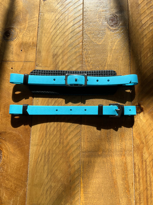 Two bright blue Hackamore Replacement Padded Nose Sets by LS Equestrian, featuring black buckles and loops, are laid flat on a textured wooden surface. Sunlight casts shadows and highlights the natural patterns in the wood.