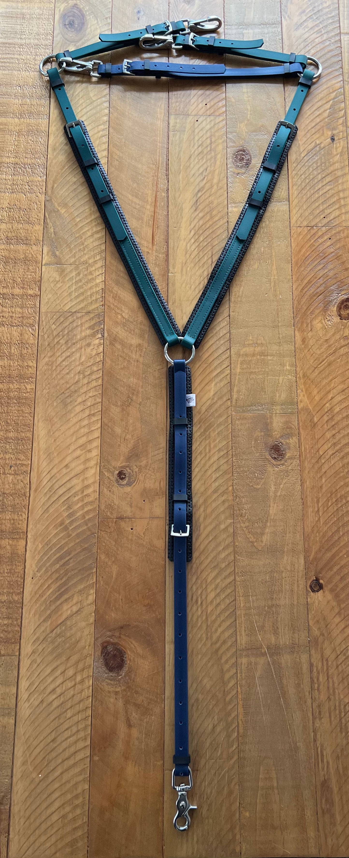 A padded breastplate from the LS Equestrian brand, in navy and hunter green, featuring metal buckles and clips, is laid out on a wooden surface. The dark green and black straps form a triangular shape, leading to a central strap equipped with adjustable buckles and a snap hook at the end.