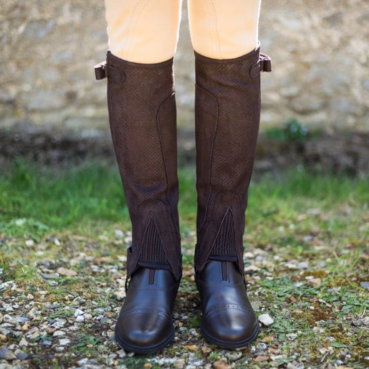 A person wearing tan riding pants and LS Equestrian's Just Chaps - Cool tall brown riding boots stands on a gravel and grassy area with a stone wall in the background. These boots, featuring reinforced toes and stirrup holders, are ideal for adventurous riding holidays.