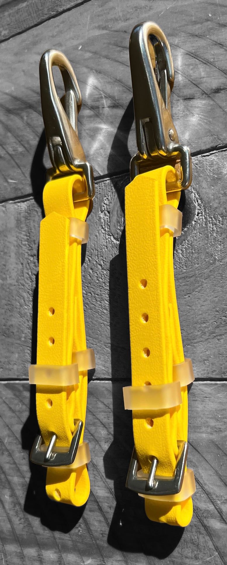Two vibrant yellow Biothane®️ Bit Hangers designed by LS Equestrian, featuring metal buckles and clasps, hang against a wooden background. Each hanger has multiple holes for adjustable sizing to ensure a secure fit.