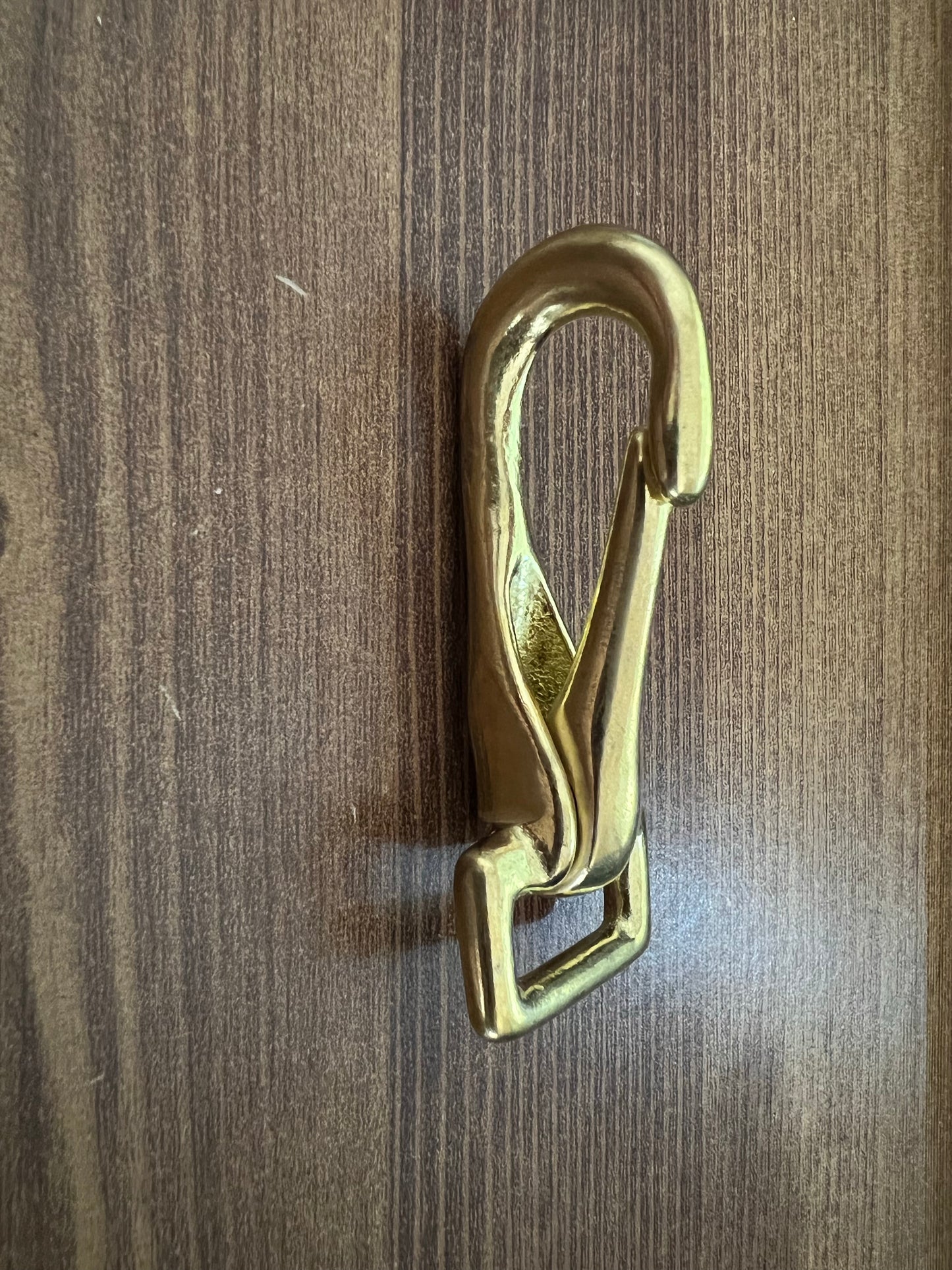 A gold-colored LS Equestrian Snap Hook / Trigger Snap with a loop at the bottom is mounted vertically against a wooden surface.