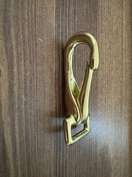 A gold-colored LS Equestrian Snap Hook / Trigger Snap with a loop at the bottom is mounted vertically against a wooden surface.