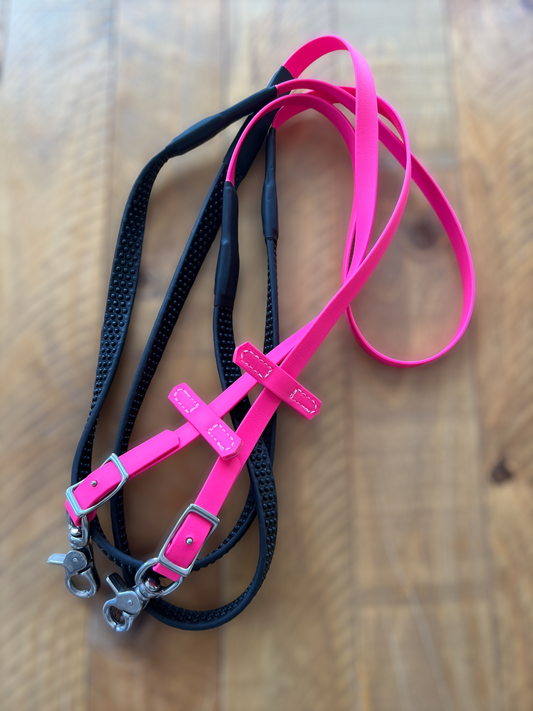 A set of dog leashes from the LS Equestrian collection is tangled on a wooden surface. One leash, named Soft Grip Non Split Reins - Hot Pink, is bright pink with silver metal clasps, while the other, custom-made, boasts black BioThane soft grip reins with textured handles.