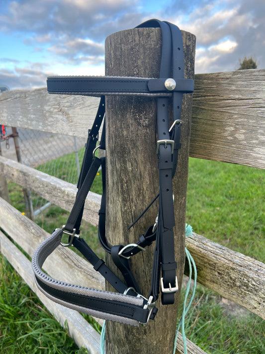 A black and grey LS Deux Bridle from LS Equestrian hangs on a wooden post beside a rustic fence, set against a grassy field and a partly cloudy sky reminiscent of an endurance riding trail.