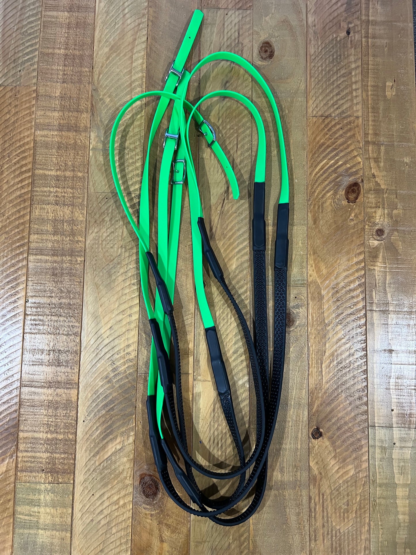 A set of LS Equestrian's Soft Grip Non Split Reins - Design your Own, customized in green and black, is laid out on a wooden floor. These reins are expertly crafted from a blend of leather and elastic materials, featuring BioThane construction with durable stainless steel fittings for added elegance and strength.