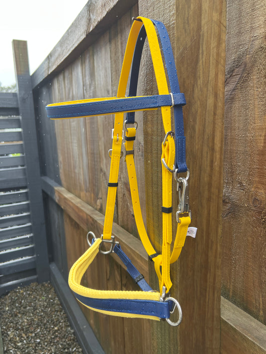 A Bitless Sidepull Bridle in Yellow & Navy from LS Equestrian is draped over a wooden fence adorned with both vertical and horizontal slats. The bridle glistens with raindrops, while pebbles scatter across the ground below.