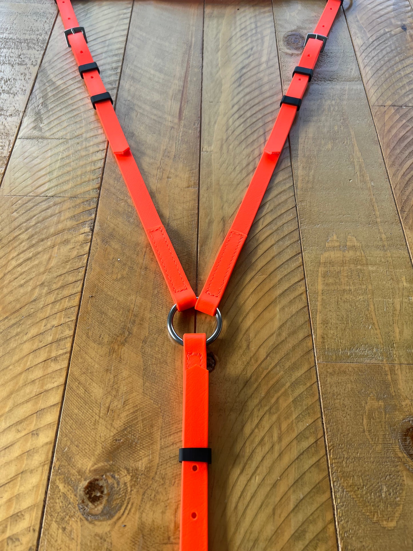 The Breastplate Zero Padding - Bright Orange from LS Equestrian is displayed on a wooden surface, showcasing its bright orange color with black accents. This custom-made design features adjustable straps and metal rings for attachments, combining style with functionality.