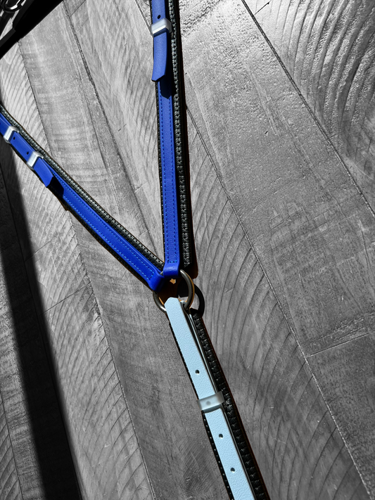 A close-up of the LS Equestrian Padded Breastplate, featuring custom-made leather straps in pastel blue and royal blue, intersecting and connected by metal rings. The breastplate is arranged on a textured gray surface, forming a geometric pattern with shadows.