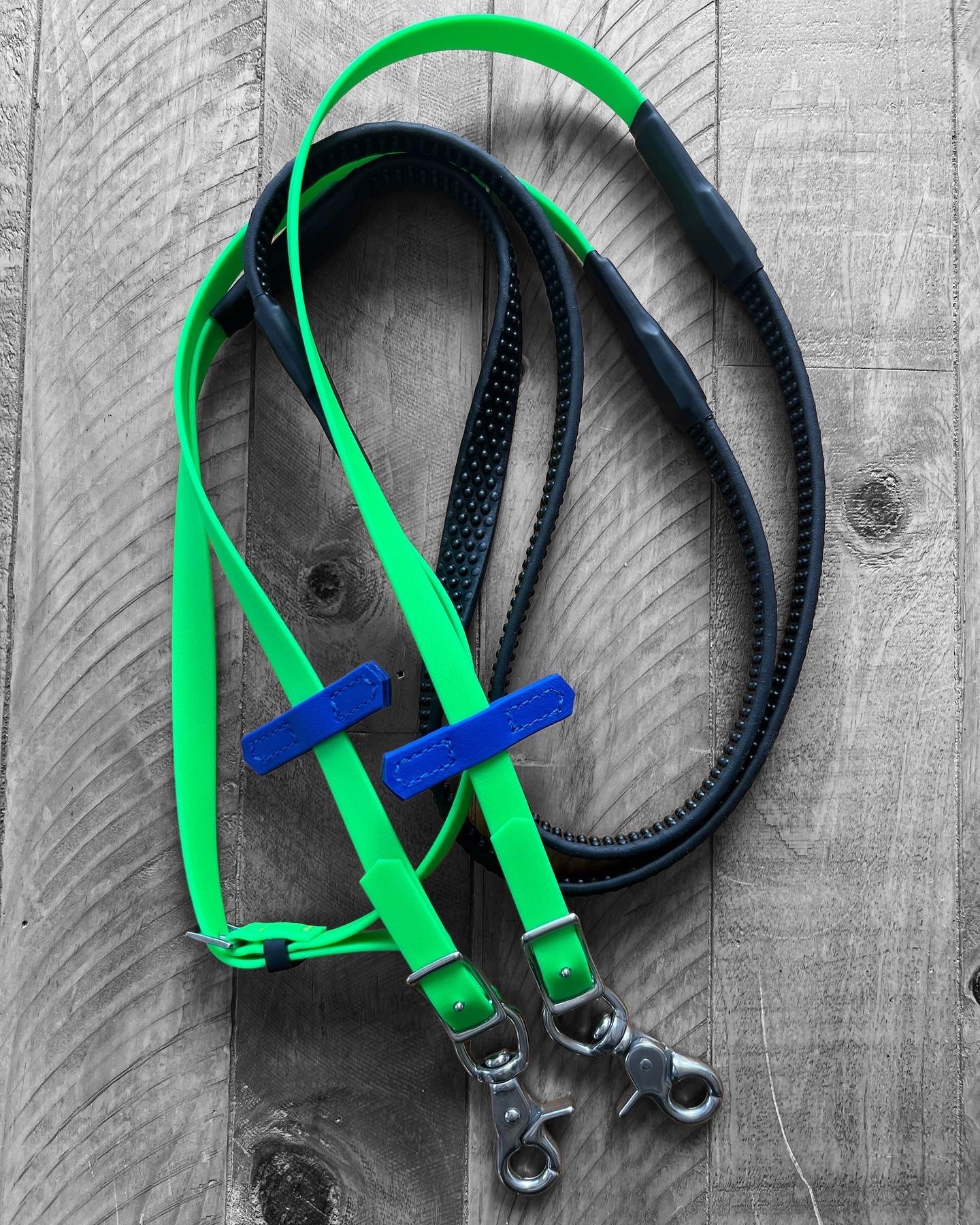 Two dog leashes in bright green and black, featuring metal clasps and blue tags, designed like the LS Equestrian Soft Grip Reins, rest elegantly on a wooden surface.