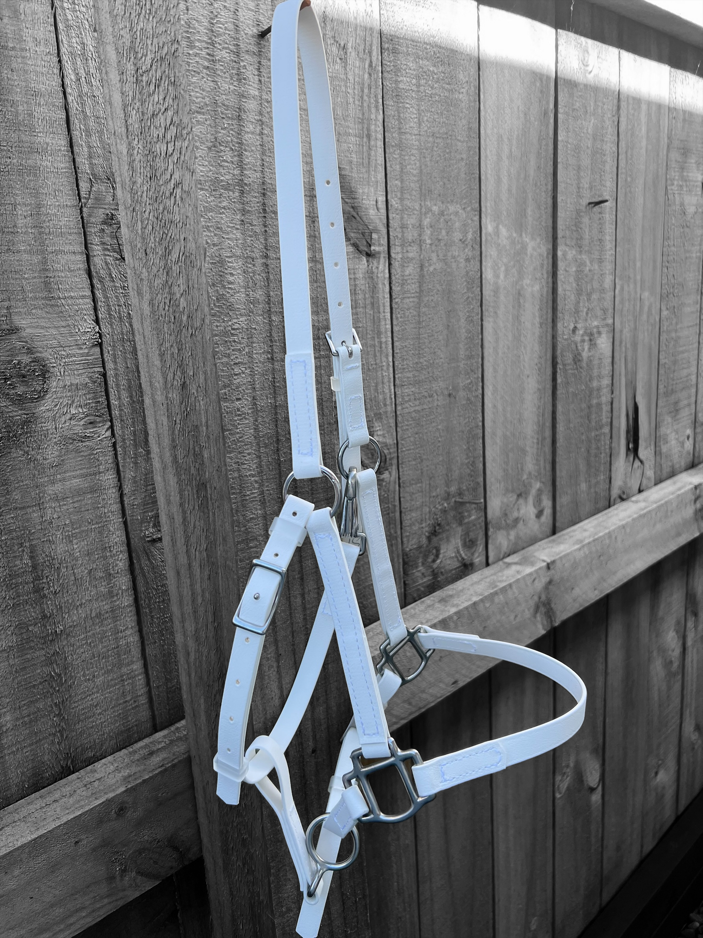 A white LS Halter from LS Equestrian is draped over a wooden fence, showcasing metal buckles and adjustable straps specifically designed for equestrian purposes. The textured wood grain of the fence highlights its custom-made quality.