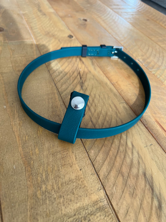A Flash Noseband with Connector from LS Equestrian, featuring teal accents, stainless steel fittings, and a metal stud with an adjustable strap, sits on a wooden surface.