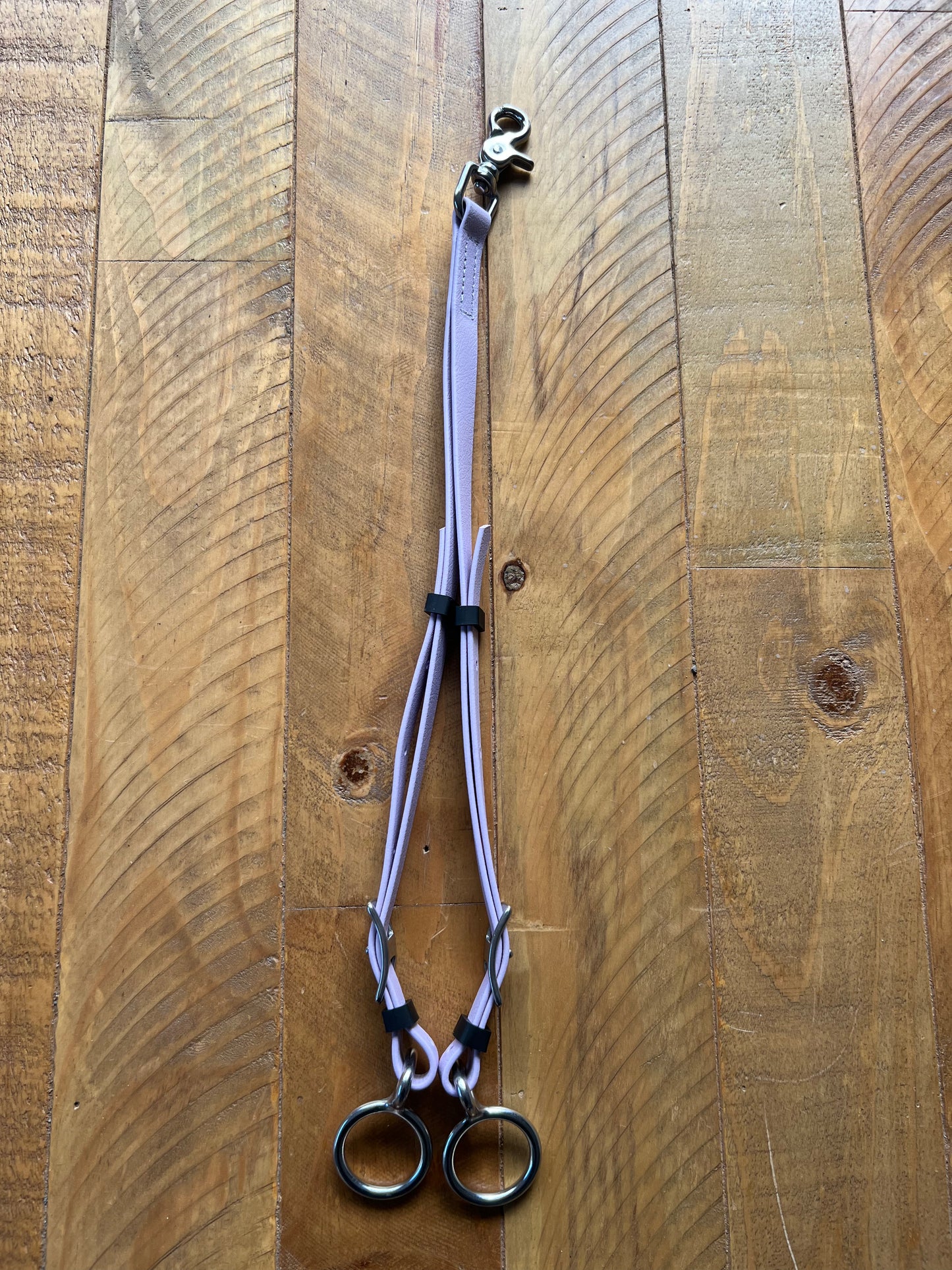 The LS Equestrian Martingale / Rings - Lavender, showcasing two pastel purple cords connected by black loops and metal clips, features a BETA finish and rests elegantly on the wooden surface.