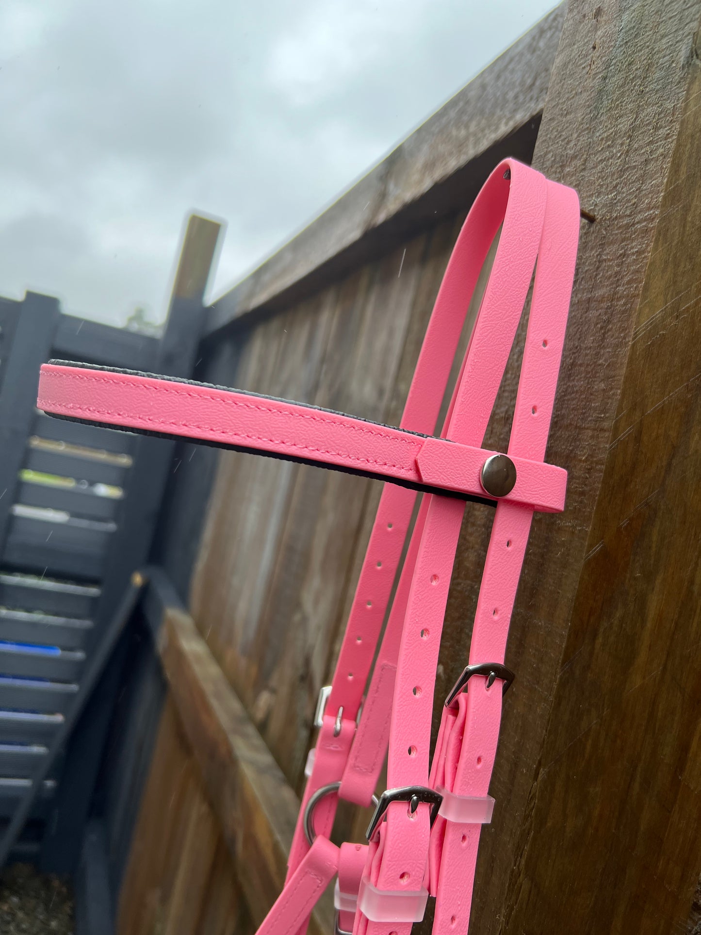 A custom-made LS Deux Bridle - Pastel Pink by LS Equestrian, featuring silver buckles, hangs gracefully on a wooden fence, glistening with moisture from a recent rain. The overcast gray sky hints at the challenges of endurance riding yet to come.