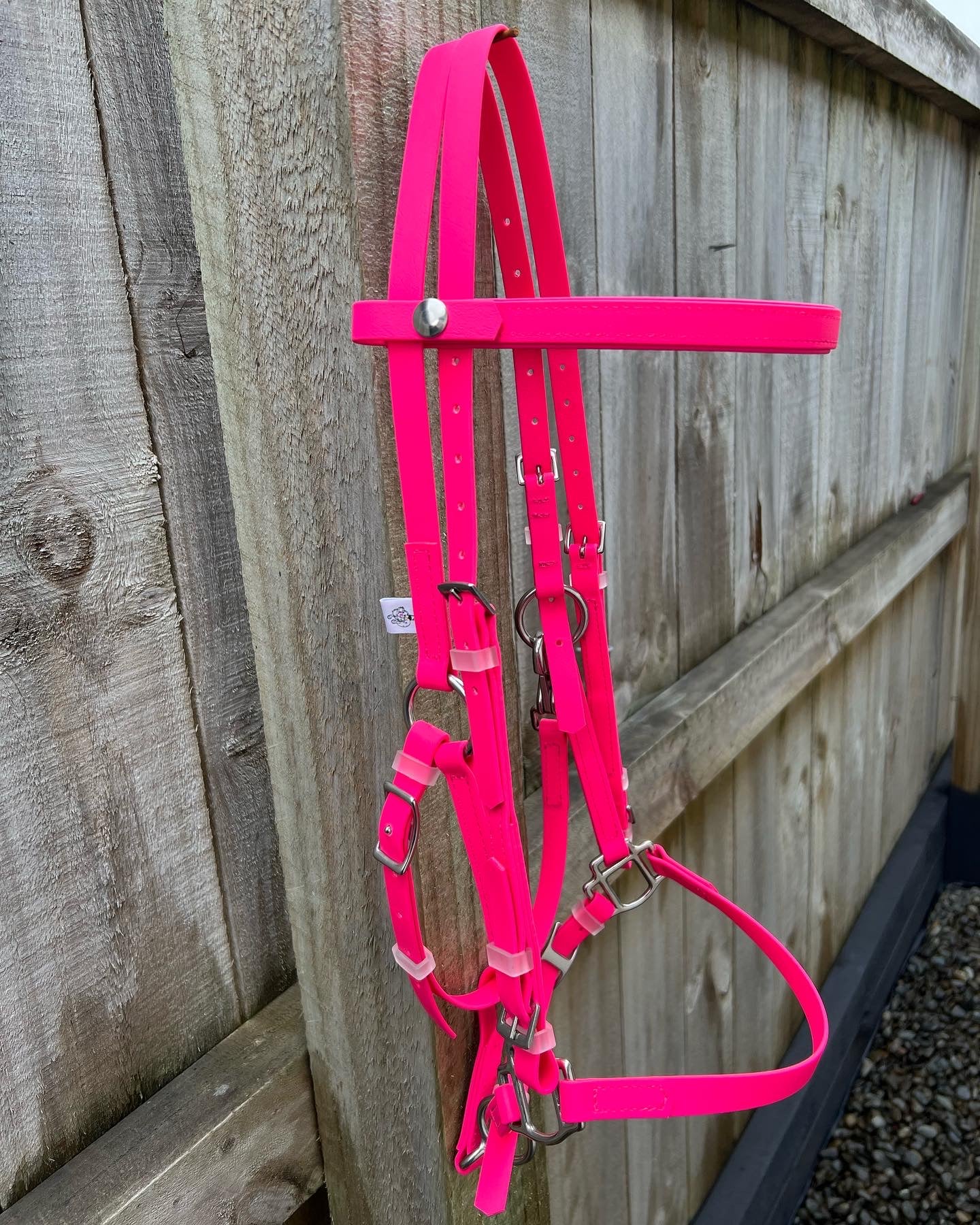 An LS Deux Bridle in hot pink from LS Equestrian with metal buckles and fittings hangs on a wooden fence. The vibrant color contrasts with the natural wood grain, showcasing its versatility as an everyday halter.