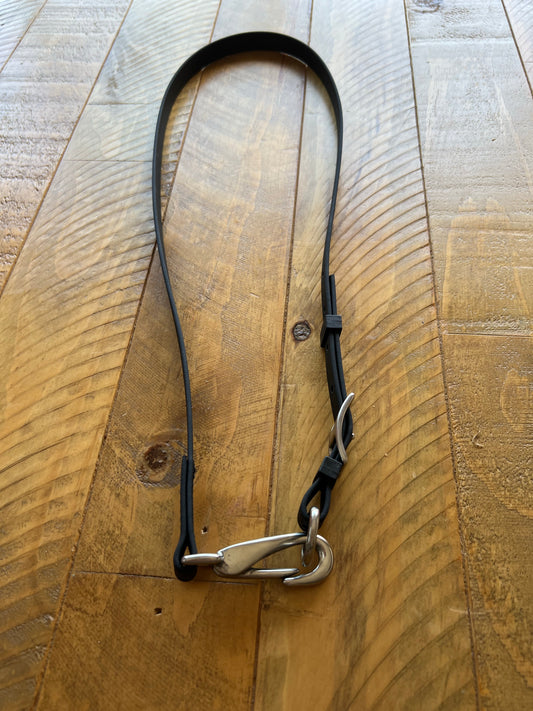 A "Design your own - Separate throatlash" by LS Equestrian, featuring a black strap with a metallic hook and loop attachment perfect for headstalls, is displayed on a textured wooden surface. This strap includes stainless steel fittings and offers adjustable loops for customization.