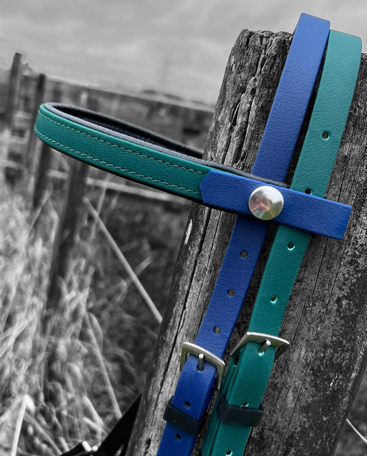 Close-up of an LS Equestrian LS Deux Bridle in padded hunter green and navy blue BioThane material, featuring metal buckles and a button, hanging on a weathered wooden post. The background shows a blurred field with a blend of black, white, and muted colors—perfect for endurance riding enthusiasts seeking durability.