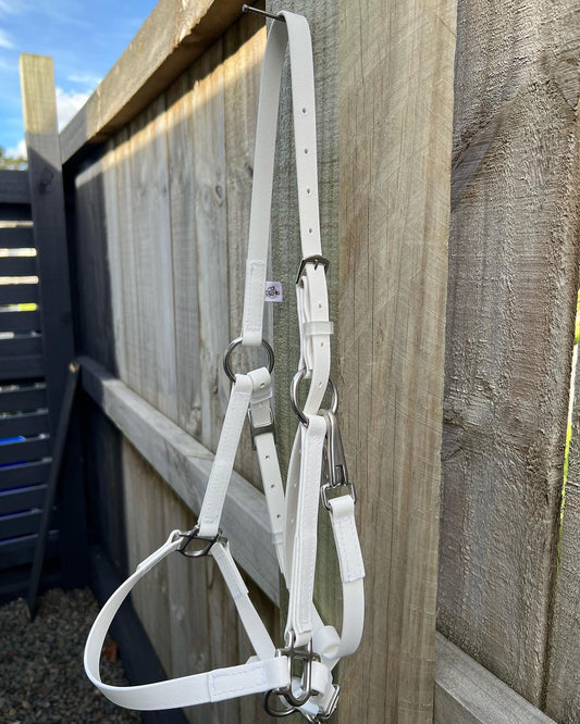 A Design Your Own - LS BioThane®️ Deux Halter from LS Equestrian in white hangs from a wooden fence. This adjustable halter, crafted from durable Beta BioThane, features silver metal buckles and hardware. The rustic weathered wood planks provide a charming backdrop, with the blue sky peeking over the neighboring fence.