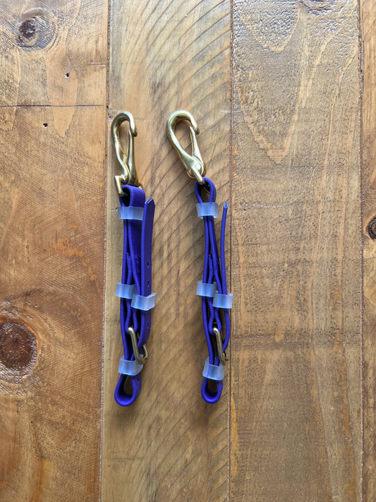 Two LS Equestrian Biothane®️ Bit Hangers, featuring gold hooks and clear plastic buckles, are crafted from Beta BioThane in a purple design and placed parallel on a wooden surface with a natural grain pattern.