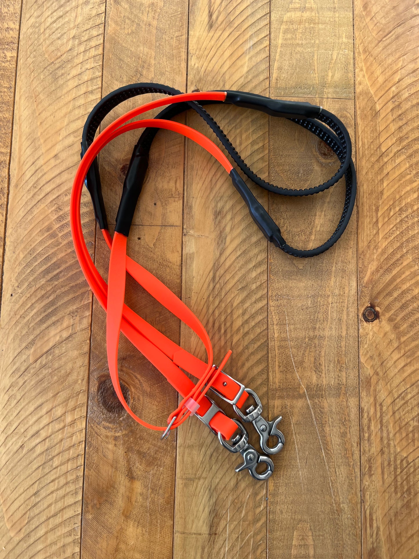 Two leashes are laid out on a wooden surface. One is from the LS Equestrian brand, featuring a soft grip in beta material with stainless steel fittings. The other leash, known as the Soft Grip Reins - Fluro Orange, boasts a vibrant hue and includes metal clasps and buckles. Both are artistically intertwined to highlight their unique design and color contrast.