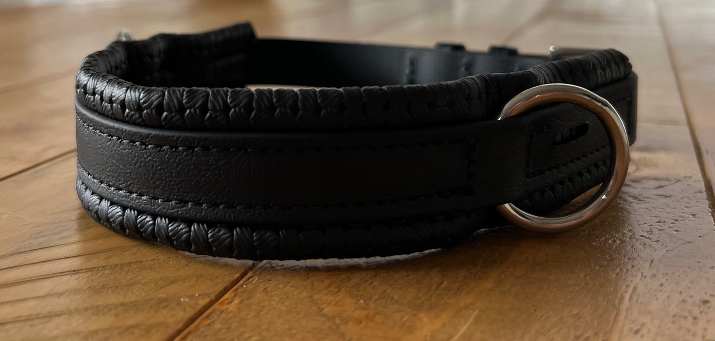 A close-up of a black Biothane®️ Sidepull Bitless Nose Convertor by LS Equestrian, showcasing its intricate craftsmanship akin to a dark leather dog collar, lies on a wooden floor. The convertor features a silver metal ring ideal for attaching a leash.
