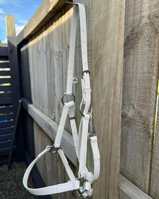 A custom-made white horse halter with sleek metallic buckles hangs on a wooden fence, set against a backdrop of wooden and metal panels. The sky peeks through softly above, reminiscent of the craftsmanship found in the LS Equestrian's LS Deux Bridle - White collection.