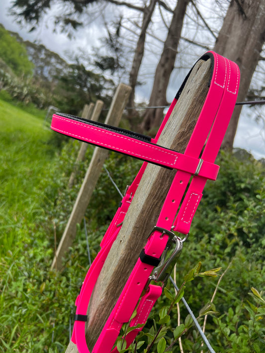 A hot pink LS Equestrian Race Bridle gracefully hangs on a wire fence outdoors, set against a blurred backdrop of trees and greenery. The vibrant color stands out against the natural environment, highlighting the custom bridle's distinct charm.